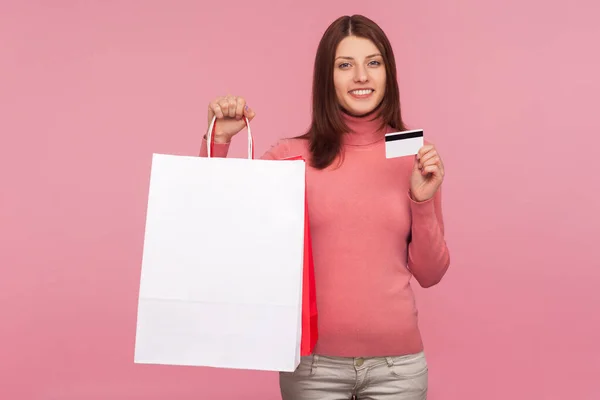 微笑积极的女人 带着棕色的头发 展示纸袋和信用卡 非接触式支付 容易用借记卡购物 在粉色背景下被隔离的室内拍摄 — 图库照片