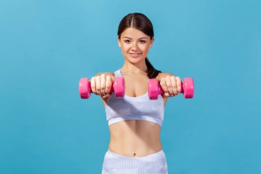 Spor kıyafetleriyle gülümseyen atletik kadınlar ellerinde dambıllar, karın kaslarını gösteriyorlar, vücudunu formda tutmak için egzersiz yapıyorlar. Mavi arka planda kapalı stüdyo çekimi