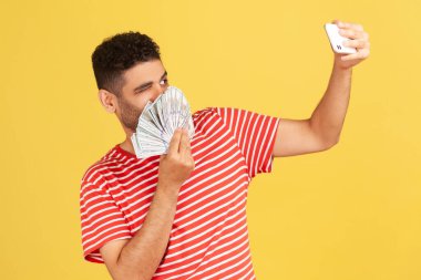 Komik zengin blogcu parayla selfie çekiyor, yüz dolarlık banknotların arkasına saklanıyor, gelirle övünüyor, bağış yapıyor. Sarı arka planda kapalı stüdyo çekimleri