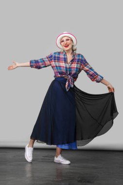 Şapka ve gözlük takan, poz veren ve kameraya mutlulukla bakan sıradan tarzda mutlu, şık, olgun bir kadının tam boy portresi. İç mekan stüdyosu gri duvar arka planında