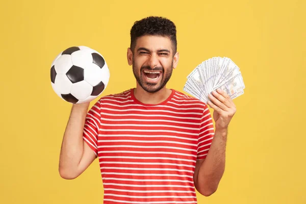 縞模様のTシャツを保持サッカーボールやドル紙幣のファン スポーツへの賭け 大きな勝利の髭を持つ満足笑顔の男 黄色の背景に隔離された屋内スタジオショット — ストック写真