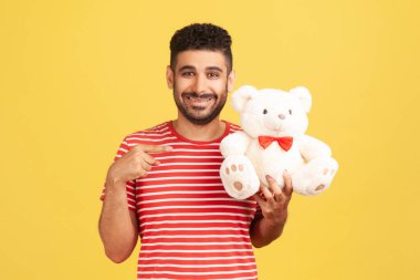 Çizgili tişörtlü, neşeli pozitif adam beyaz pofuduk ayıyı işaret ederek sana romantik bir hediye hazırlıyor. Sarı arka planda kapalı stüdyo çekimleri
