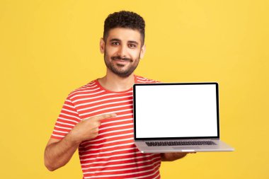 Çizgili tişörtlü, sakallı, boş beyaz ekranlı dizüstü bilgisayarı işaret eden, online eğitim dersleri öneren olumlu bir adam. Sarı arka planda kapalı stüdyo çekimleri