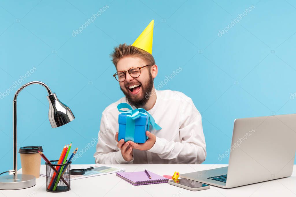 Happy positive man office worker with beard in white shirt and cone hat holding gift box and laughing, sitting at workplace with laptop enjoying present. Indoor studio shot isolated on blue background