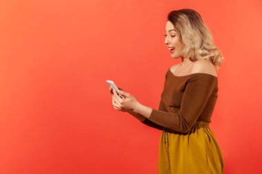 Yan görünüş, akıllı telefon tutan, hoş mesajlar okuyan, e-posta atan ve gülümseyen mutlu bir kadın. Turuncu arka planda kapalı stüdyo çekimleri