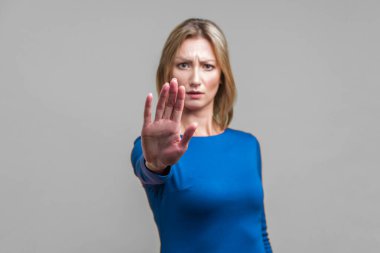 Hayır, yasak! Dar mavi elbiseli üzgün genç bir kadının portresi. Durma hareketi, içki yasağı veya endişeli suratlı uyarı ifadesi sergiliyor. Gri arka planda kapalı stüdyo çekimi