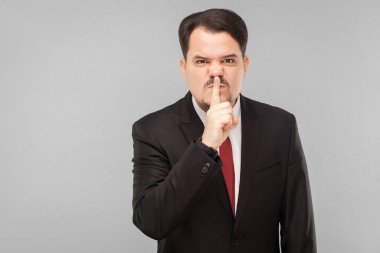 Susun! Toplantı var, sakın ses çıkarmayın. Kapalı stüdyo çekimi. Gri arka planda izole. Siyah takım elbiseli, kırmızı kravatlı ve bıyıklı yakışıklı işadamı kızgın yüzlü kameraya bakıyor..