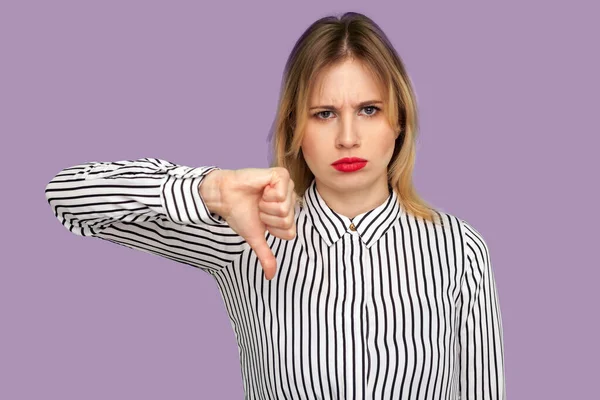 Dislike! Attractive blond woman frowning with naughty expression and showing thumbs down, expressing disapproval, negative feedback. indoor studio shot isolated on purple background