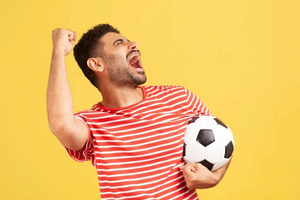 ストライプの赤いTシャツを着た非常に肯定的な満足した男は チャンピオンシップで好きなサッカーチームの勝利を祝う サッカーボールを保持叫んでいます 黄色の背景に隔離された屋内スタジオショット — ストック写真