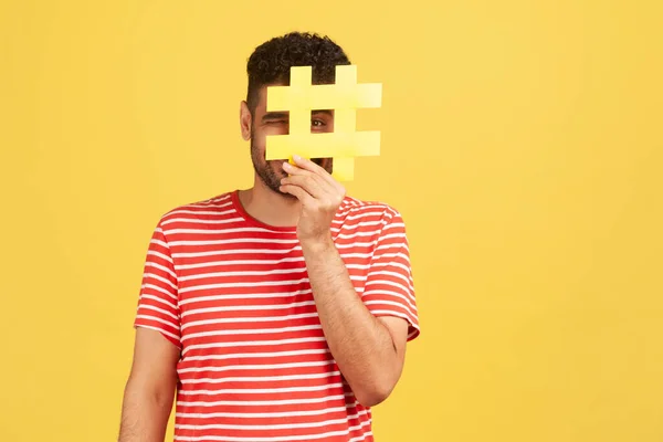 黄色のハッシュタグ記号の後ろに顔を隠す縞模様のTシャツの面白い男ブロガーやSmmマネージャ ウェブサイト上のトピックをタグ付けするために教える 推奨事項を与える 黄色の背景に隔離された屋内スタジオショット — ストック写真