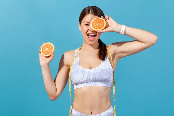 Smiling active woman in white sportswear with tape line on shoulders closing eye with grapefruit half, having fun and losing extra weight on fruits diet. Indoor studio shot isolated on blue background