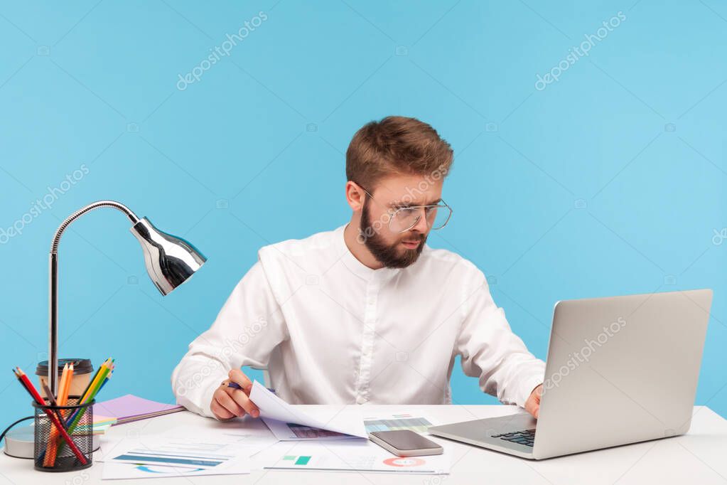 Concentrated man analyst in eyeglasses and white shirt checking data on graphs, inserting results at laptop program, sitting at workplace. Indoor studio shot isolated on blue background.