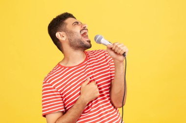 Çizgili tişörtlü olumlu sakallı adam yüksek sesle şarkı söylüyor mikrofonu elinde tutuyor, karaoke yaparken dinleniyor, şarkıcı performansı sergiliyor. Sarı arka planda kapalı stüdyo çekimleri