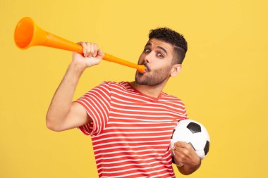 Kırmızı çizgili tişörtlü heyecanlı sakallı adam elinde siyah beyaz futbol topuyla boynuz sallayarak favori futbol takımının zaferini kutluyor. Sarı arka planda kapalı stüdyo çekimleri