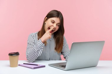 Tembel kadın ofis çalışanı burun karıştırırken ve dikkatsiz bir ifadeyle laptop ekranına bakarken iş yerindeki video konuşmasında eğleniyor. Pembe arka planda kapalı stüdyo çekimleri