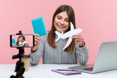 Akıllı telefon kamerasında pasaport ve kağıt uçak gösteren olumlu kadın blogcu, turla övünen, tatile çıkan. Pembe arka planda kapalı stüdyo çekimleri