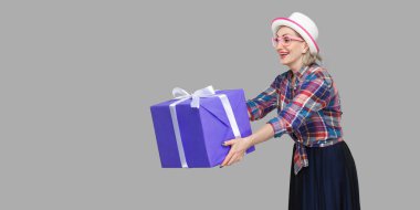 Beyaz şapkalı, kareli gömlek standı giymiş, dişlek gülüşlü büyük hediye kutusunu paylaşan mutlu, süslü büyükannenin yan görünümü. Gri arka planda kapalı stüdyo çekimi.