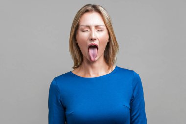 Portrait of positive carefree woman with fair hair in blue dress standing with closed eyes and demonstrating tongue, naughty disobedient expression. indoor studio shot isolated on gray background clipart