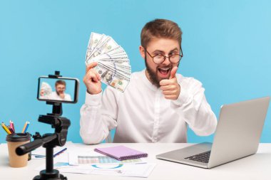 Gözlüklü ve tişörtlü mutlu olumlu adam blogcusu bir dolar nakit tutuyor akıllı telefon kamerasında baş parmaklarını gösteriyor, video kaydediyor, kazanç dersi veriyor. Mavi arka planda kapalı stüdyo çekimi