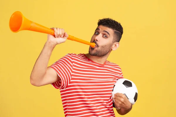 黒と白のサッカーボールを保持ホーンに吹いて赤い縞模様のTシャツで興奮髭の男は お気に入りのサッカーチームの勝利を祝う 黄色の背景に隔離された屋内スタジオショット — ストック写真