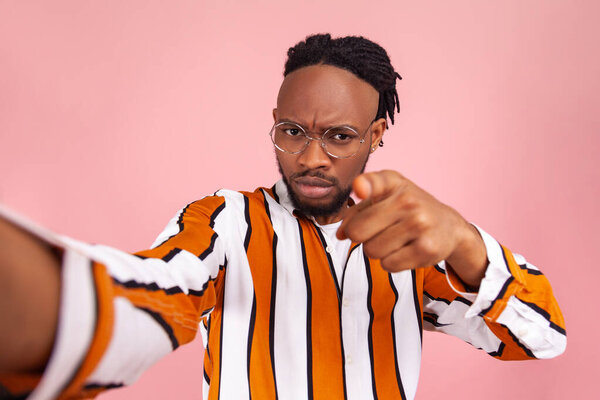 Hey you! Strict rude afro-american man blogger with beard and dreadlocks in striped shirt pointing finger at you seriously looking at selfie camera. Indoor studio shot isolated on pink background