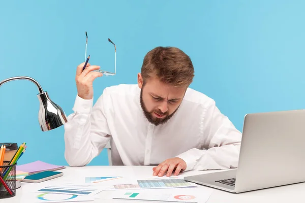 专心致志的大胡子男人放下眼镜 在统计报告中用心学习信息 用笔记本电脑分析工作场所的数据 室内拍摄被蓝色背景隔离 — 图库照片