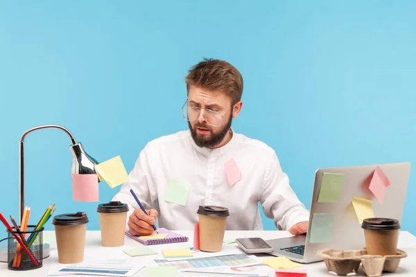 職場に座っている忙しい男のオフィスワーカーは すべて粘着性のあるメモで覆われ メモ帳に思考を書き留め コーヒーを飲んでいます 青い背景に隔離された屋内スタジオショット — ストック写真