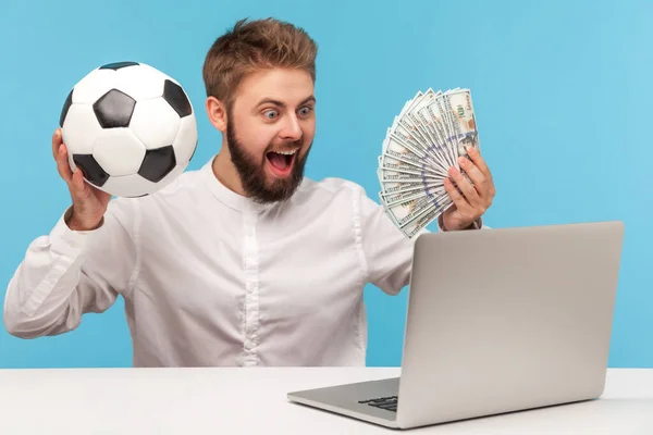 ドルの現金とラップトップディスプレイを見てサッカーボールのファンを保持髭を持つ幸せな興奮した男 賭けとジャックポットを獲得 青い背景に隔離された屋内スタジオショット — ストック写真