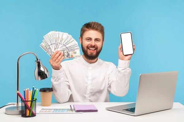 一个面带微笑 留着胡子 穿着白衬衫的男人 他展示了大量的100美元钞票和白屏智能手机 银行和网上应用的信用记录 室内拍摄被蓝色背景隔离 — 图库照片