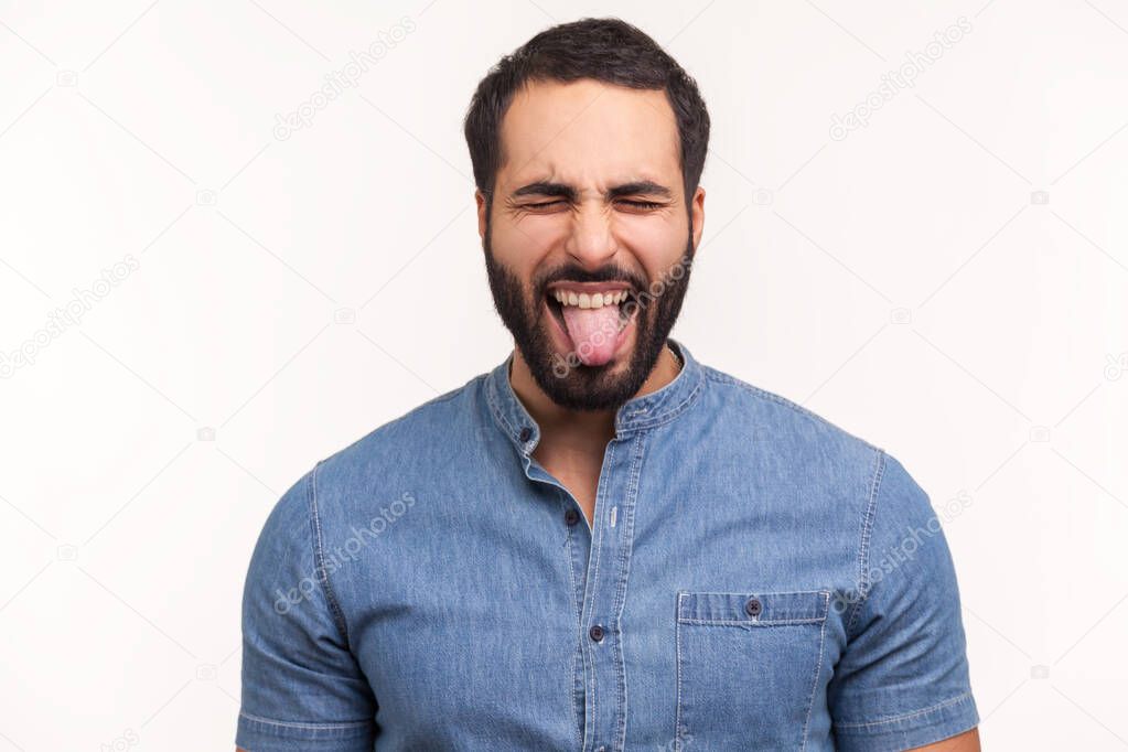 Naughty infantile man with beard closing eyes and showing tongue, having fun, fooling around, childish manners. Indoor studio shot isolated on white background