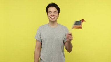 Amerika Birleşik Devletleri bayrağını sallayan gri tişörtlü, vatansever genç adam dişlek tebessümle kameraya bakıyor, seçimler, bağımsızlık günü. Sarı arka planda kapalı stüdyo çekimleri