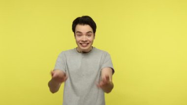 Gel kollarıma! Keyifli, iyimser, gri tişörtlü genç bir adam. Seni arıyor, elini uzatıyor, dostça gülümsüyor ve sarılıyor. Sarı arka planda kapalı stüdyo çekimleri