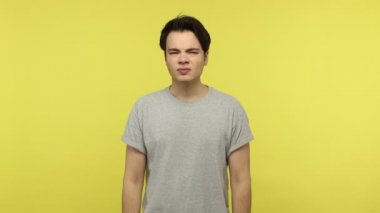 Diş sorunları. Gri tişörtlü sağlıksız genç adam ağrısından çatık kaşlı yanağa dokunuyor diş ağrısı, çürük ya da diş eti iltihabı belirtileri gösteriyor. Sarı arka planda kapalı stüdyo çekimleri