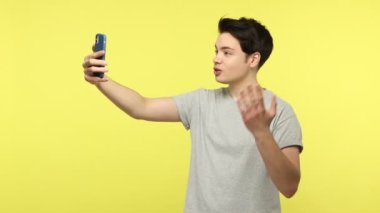 Gri tişörtlü neşeli genç blog yazarı, takipçileriyle akıllı telefondan iletişim kuruyor, yeni gönderisini ve aboneliğini derecelendirmek istiyor. Sarı arka planda kapalı stüdyo çekimleri