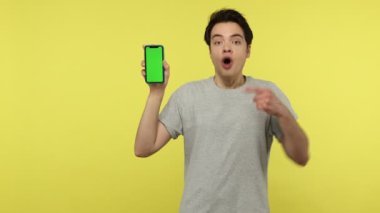 Gri tişörtlü aşırı mutlu genç yeşil ekran akıllı telefonunda uyguladığı havalı uygulamayla şok oldu ve baş parmağını gösteren kameraya baktı. Sarı arka planda kapalı stüdyo çekimleri