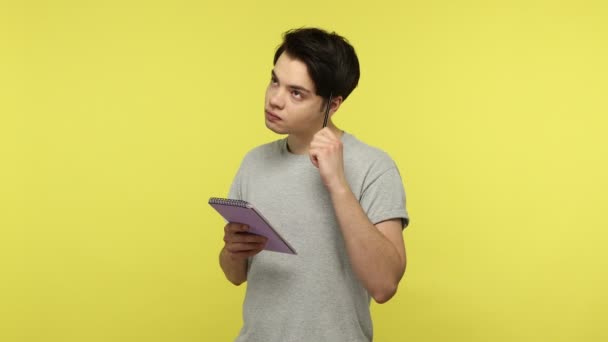 有思想的年轻人穿着休闲风格的灰色T恤 站在那里 用铅笔刻划着沉思的表情 把想法写在笔记本上 在黄色背景下隔离的室内拍摄 — 图库视频影像