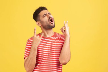 Çizgili tişörtlü itaatsiz yaramaz adam rock 'n roll hareketi gösteriyor ve heyecanla bağırıyor, eğleniyor, ağır müzikten zevk alıyor. Sarı arka planda kapalı stüdyo çekimleri