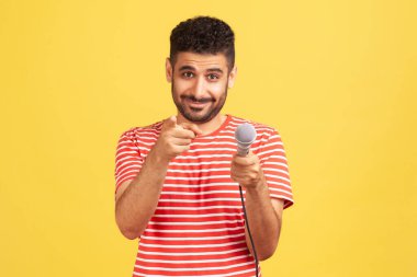Çizgili kırmızı tişörtlü, gülümseyen sakallı adam mikrofon tutuyor ve kamerayı işaret ediyor. Şarkı söylemeni ya da tebrik etmeni istiyor. Sarı arka planda kapalı stüdyo çekimleri