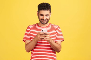 Çizgili tişörtlü mutlu adam akıllı telefonda mesajlaşıyor, sosyal ağlarda sohbet ediyor ya da kuryeyle çevrimiçi sohbet ediyor. Sarı arka planda kapalı stüdyo çekimleri