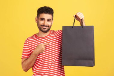 Çizgili tişörtlü, gülümseyen sakallı adam alışverişle, mevsimlik satışla tatmin olan siyah kağıt torbayı işaret ediyor. Sarı arka planda kapalı stüdyo çekimleri