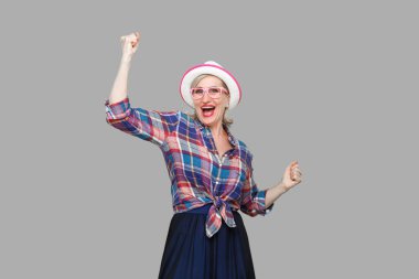 Mutlu, modern stil sahibi olgun bir kadının portresi şapkası ve gözlükleriyle ayakta durup zaferini kutluyor ve kameraya bakıyor. Stüdyo çekimi gri arkaplanda izole edildi.