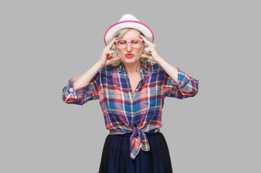 Baş ağrısı veya kafa karışıklığı. Somurtkan, olgun bir kadının portresi. Sıradan bir tarzda. Şapkası ve gözlükleriyle başını baş ağrısından tutuyor. Gri arka planda kapalı stüdyo çekimi.