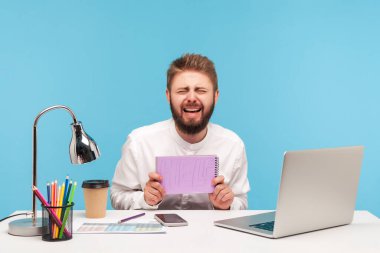 Yorgun adam ofis çalışanı not defterinde yazılı yardım ve ağlama belirtileri gösteriyor. Oturma yerinde oturuyor. Ekstra çalışmaktan ve hafta sonları vardiyalardan, profesyonel yorgunluktan bıkmış. Mavi arka planda kapalı stüdyo çekimi