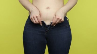 Yakından şişman kadın kot pantolon giyiyor, şişman göbeğinin düğmesini kapatmaya çalışıyor, kilo verme problemleri, zayıf ve sağlıklı olma arzusu. Sarı arka planda kapalı stüdyo çekimleri