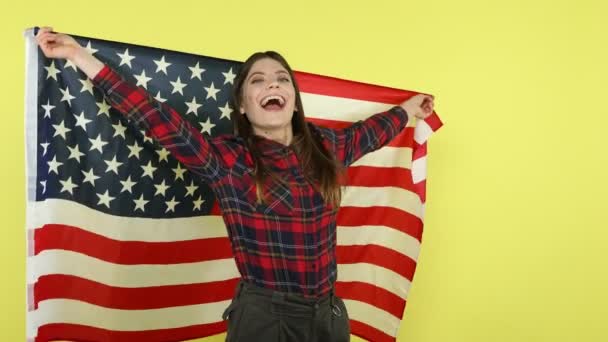 チェック柄のシャツを着た幸せな愛国的な女性がアメリカ国旗を包み 独立記念日 選挙を笑顔で祝います 黄色の背景に隔離された屋内スタジオショット — ストック動画