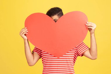 Kırmızı çizgili tişörtlü ürkek bir adam utangaç bir şekilde büyük kırmızı kağıt kalpten dışarı bakıyor, duygularını gösteriyor, sevgililer gününde sürpriz yapıyor. Sarı arka planda kapalı stüdyo çekimleri