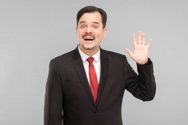 Hey, merhaba! Pozitif patron merhaba işareti gösteriyor, dişlek kameraya gülümsüyor, astlarını gördüğüne seviniyor. Kapalı, gri arka planda izole edilmiş. Siyah takım elbiseli, kırmızı kravatlı ve bıyıklı yakışıklı bir iş adamı..