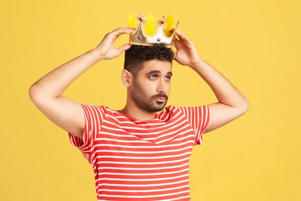 Ambitious bearded man wearing golden crown, imagining promotion at work to position of top manager or boss, looking with arrogance, privileged status. Indoor studio shot isolated on yellow background