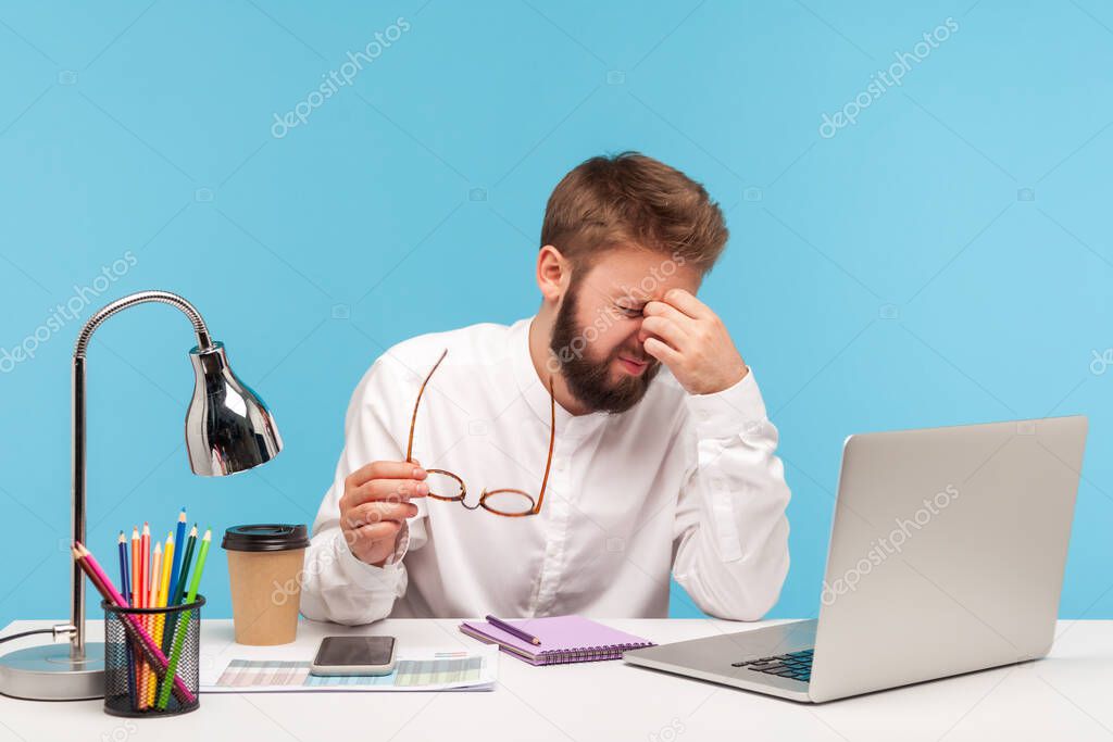 Exhausted stressed man office worker rubbing tired of laptop work eyes, putting off glasses, sitting workplace, overworking, problems with vision. Indoor studio shot isolated on blue background