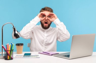 Şok olmuş sakallı bir ofis çalışanı iş yerinde dizüstü bilgisayarla oturuyor, dürbünle el hareketleriyle ağzını açıyor, büyük bir şaşkınlık ve şaşkınlık içinde. Mavi arka planda kapalı stüdyo çekimi